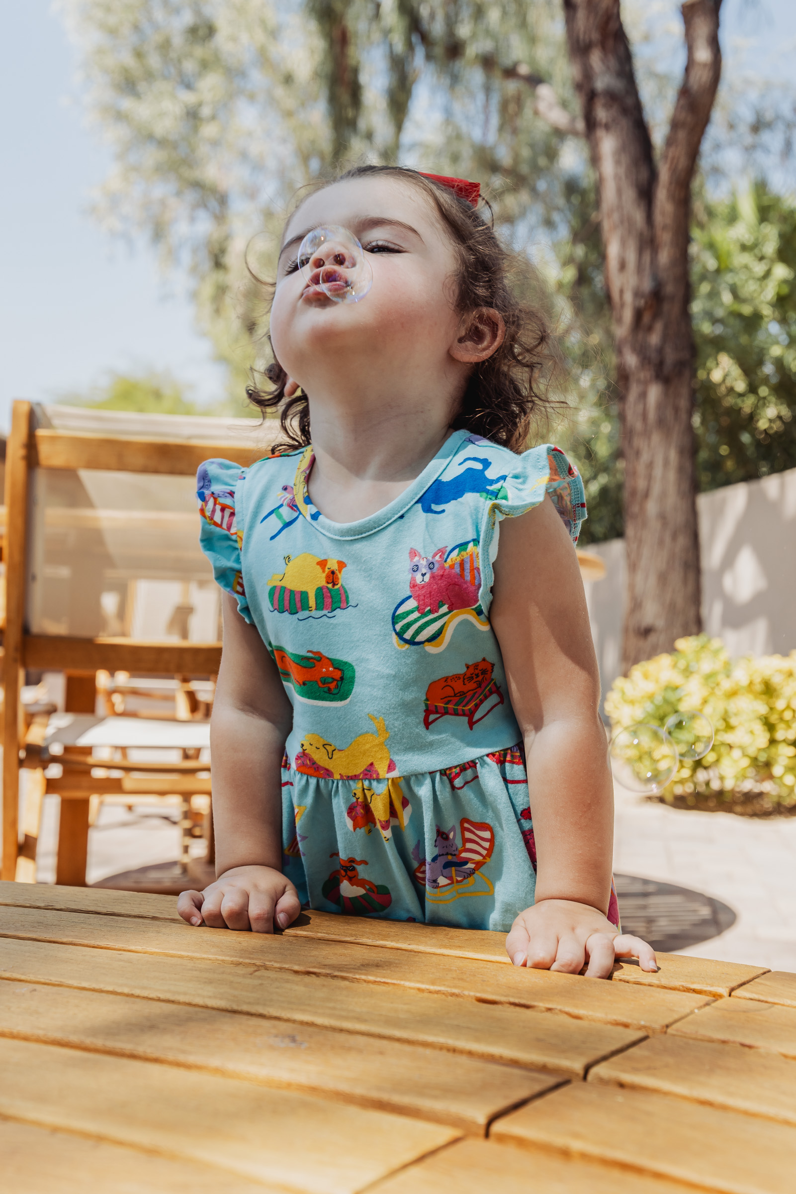 Toddler girl in a patterned dress making a playful puckered-lip face while leaning on a wooden outdoor table