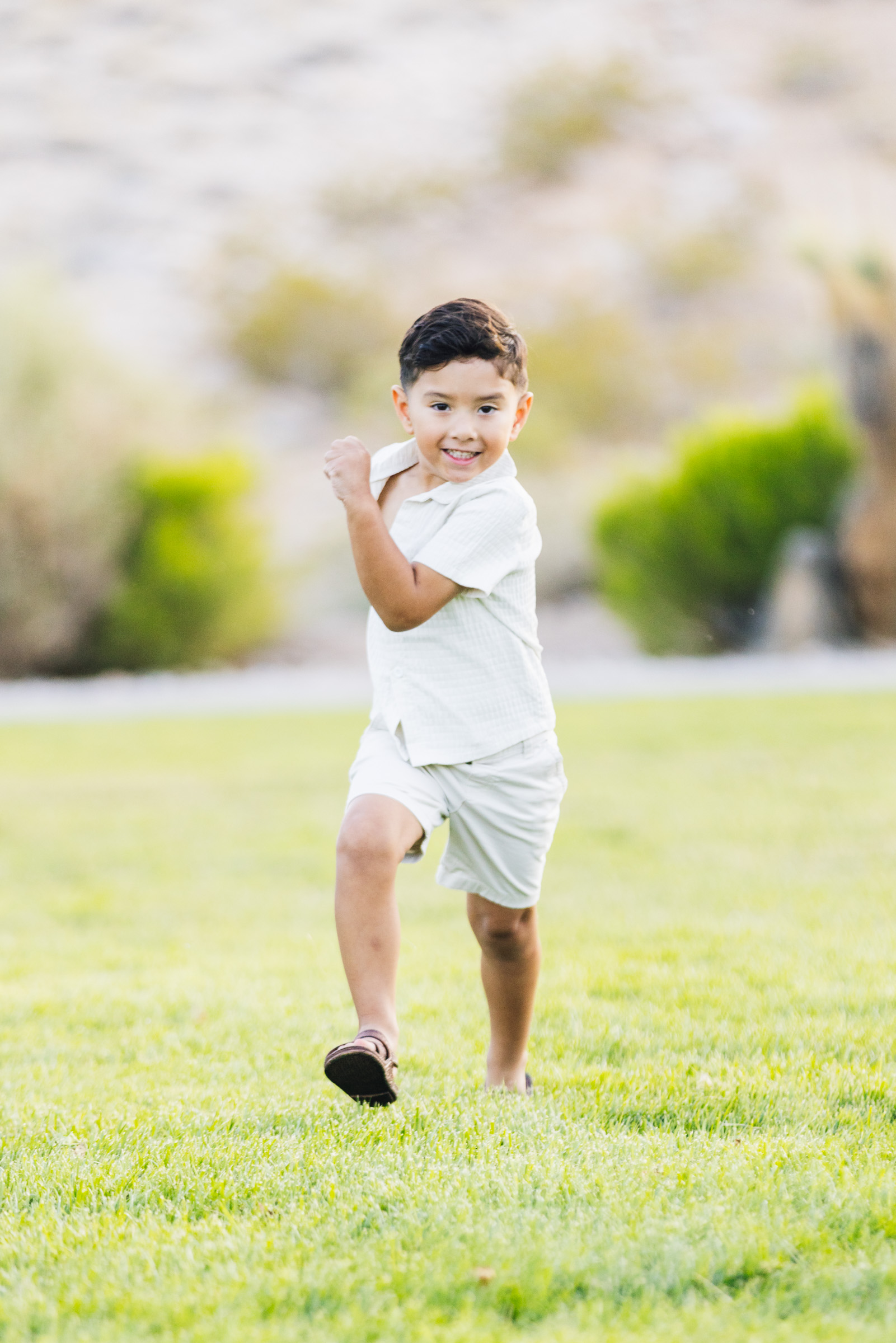 Young boy in a casual white outfit running toward the camera across the grass with a big smile