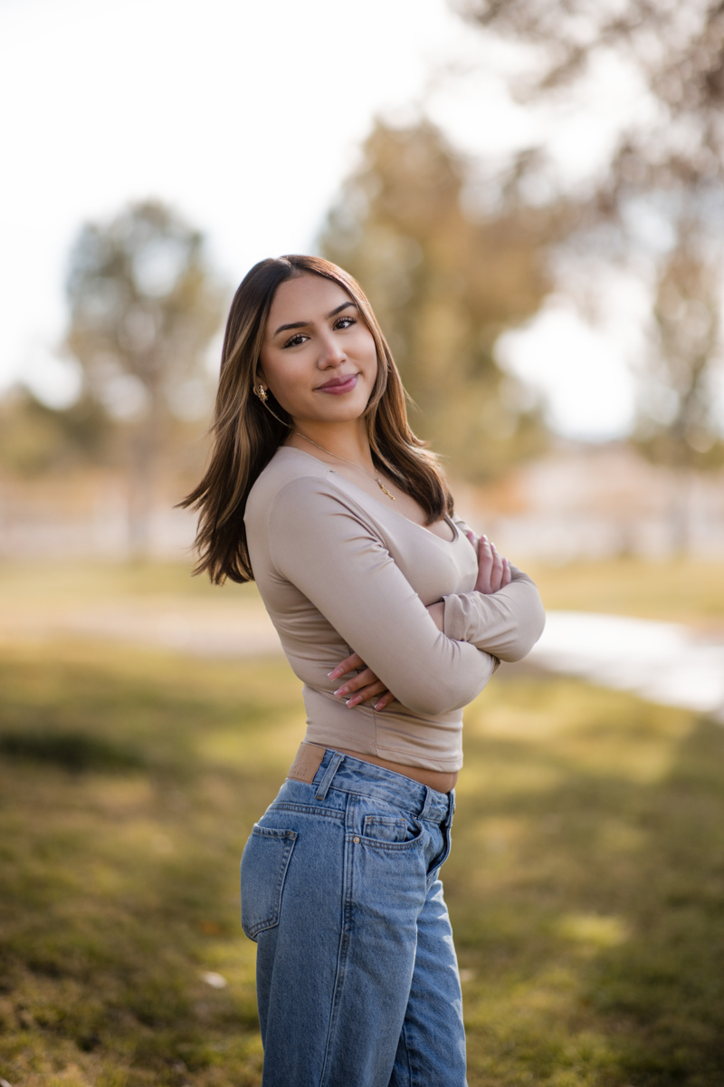 This outdoor portrait session image, photographed in Las Vegas by Joseph Blake, features a young woman standing with arms crossed against a blurred background of trees after retouching.