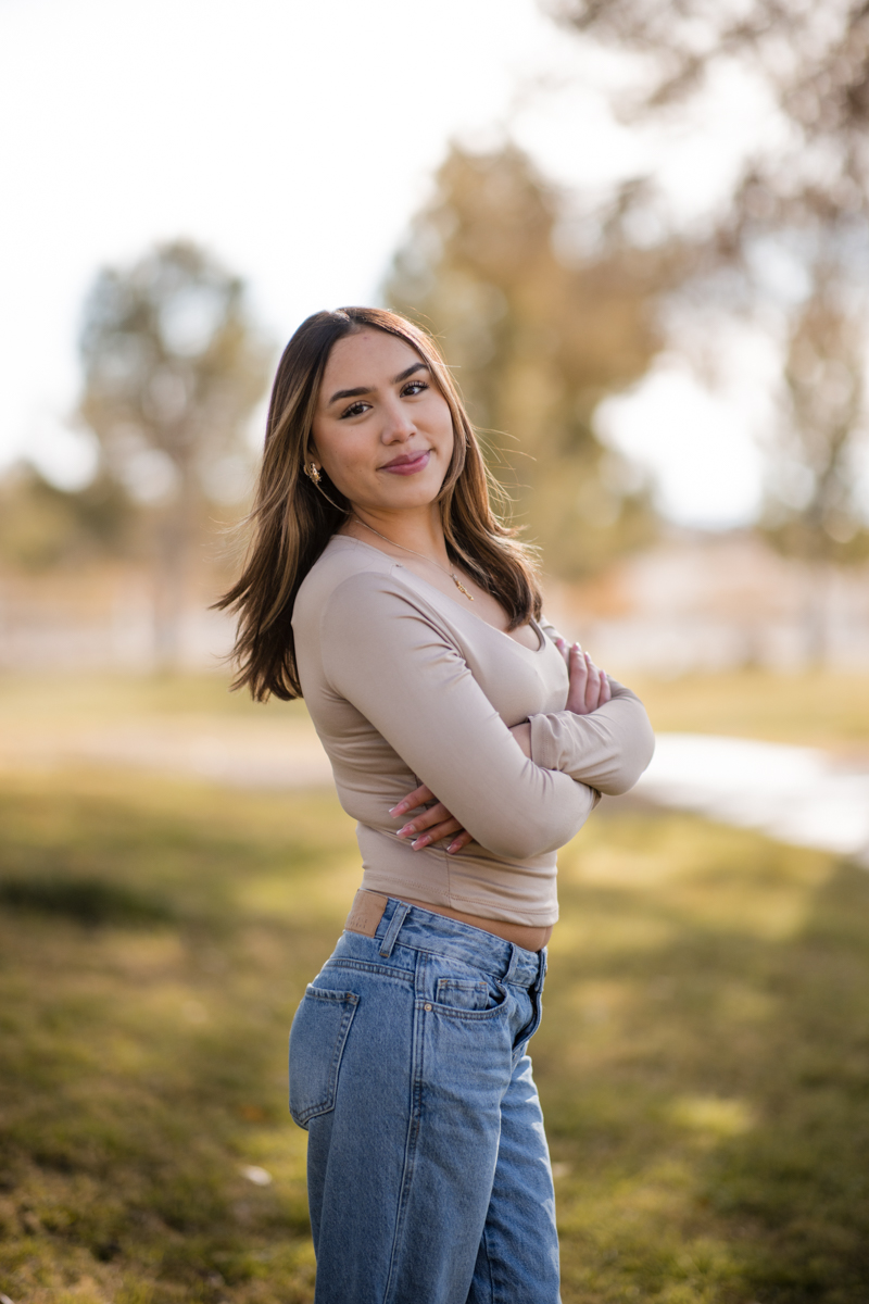 This outdoor portrait session image, photographed in Las Vegas by Joseph Blake, features a young woman standing with arms crossed against a blurred background of trees before retouching.